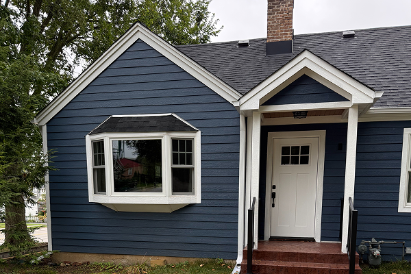Exterior view of a dark blue siding home featuring a newly installed white bay window on the left and a white front entry door with a covered porch.
